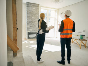 Two construction professionals, one in a hard hat, examine plans in a sunlit, unfinished room with a fireplace and stone wall.