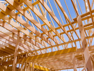 Wooden roof trusses under construction against a clear blue sky. Sunlight casts shadows, highlighting the natural wood texture.