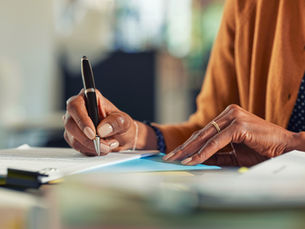 Close-up of a person’s hands signing a document with a pen at a desk, with papers, folders, and office supplies in the background.