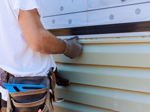 Worker in a white shirt and gloves installs light-colored siding on a house. Tool belt with a blue square visible. Focused effort.