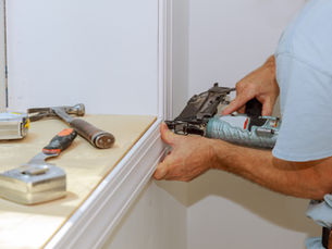 Person using a nail gun on white trim indoors. Nearby tools include a tape measure, hammer, and paint scraper on a beige surface.