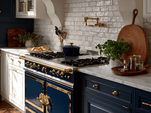 Luxurious kitchen with a navy and gold stove, marble countertops, white tile backsplash, fresh bread, herbs, and spices on display.