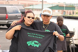 Two people are smiling and posing together in a parking lot. The person on the left is wearing sunglasses and has their arm around the other person, who is wearing a "Brown N Proud" cap. Both individuals are holding up a black T-shirt with green graphics of a car and the text "Return Of The Funk Kickback Hosted By Ashley Garcia." Other people and vehicles are visible in the background.
