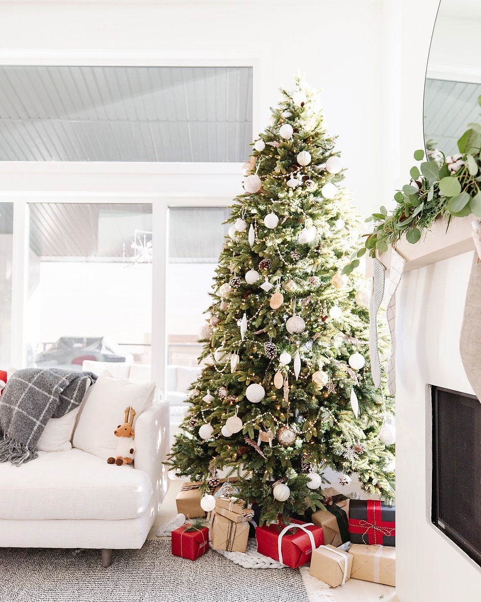 White lights Christmas tree white glass ornaments reindeer acorn glass bead garland hand wrapped paper presents under tree white sofa reindeer stuffed animal grey plaid throw blanket natural light fireplace with eucalyptus garland