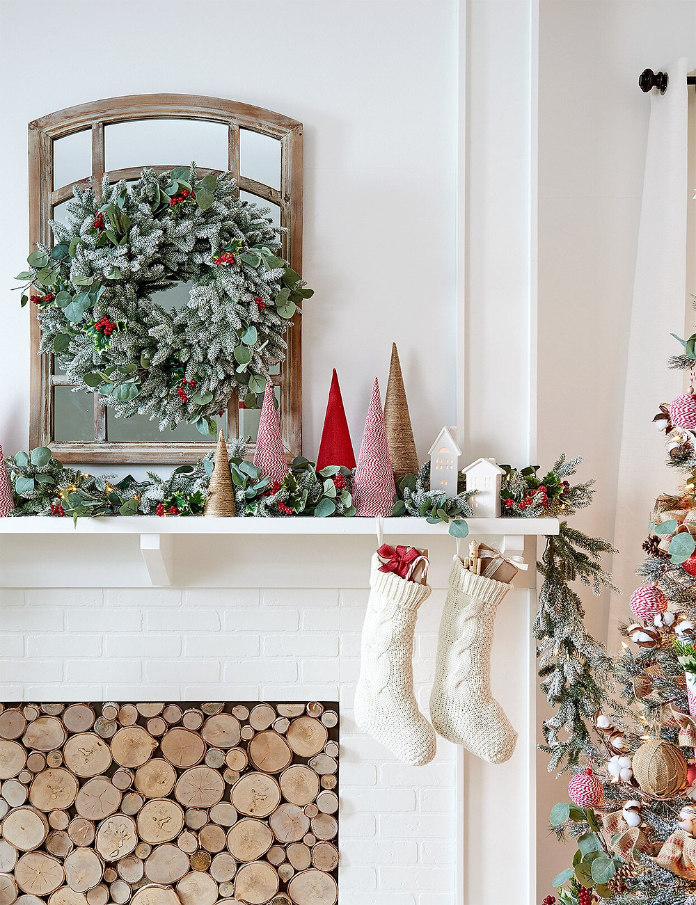 A white Christmas holiday fireplace with a mantel decorated with greenery, a mirror, bottle brush trees and stockings for a stylish and modern look.