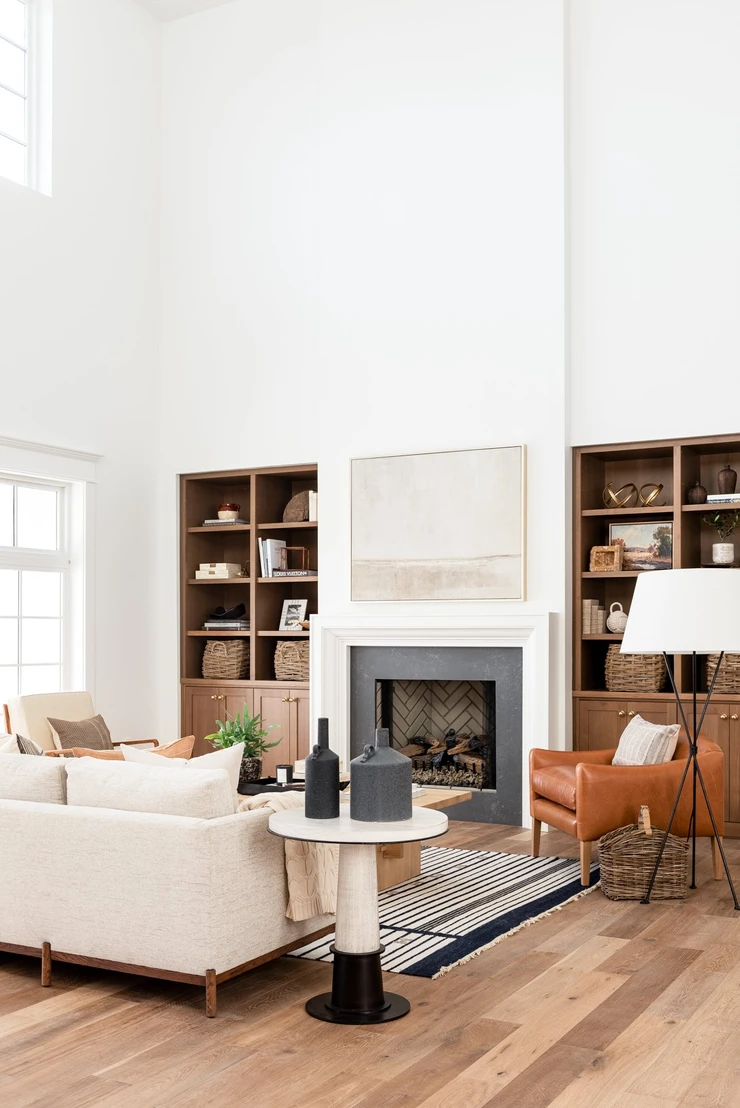 A modern farmhouse living room with a tall white fireplace with a black surround next to some natural wood built in shelves and a white cream sofa with a caramel leather chair. 