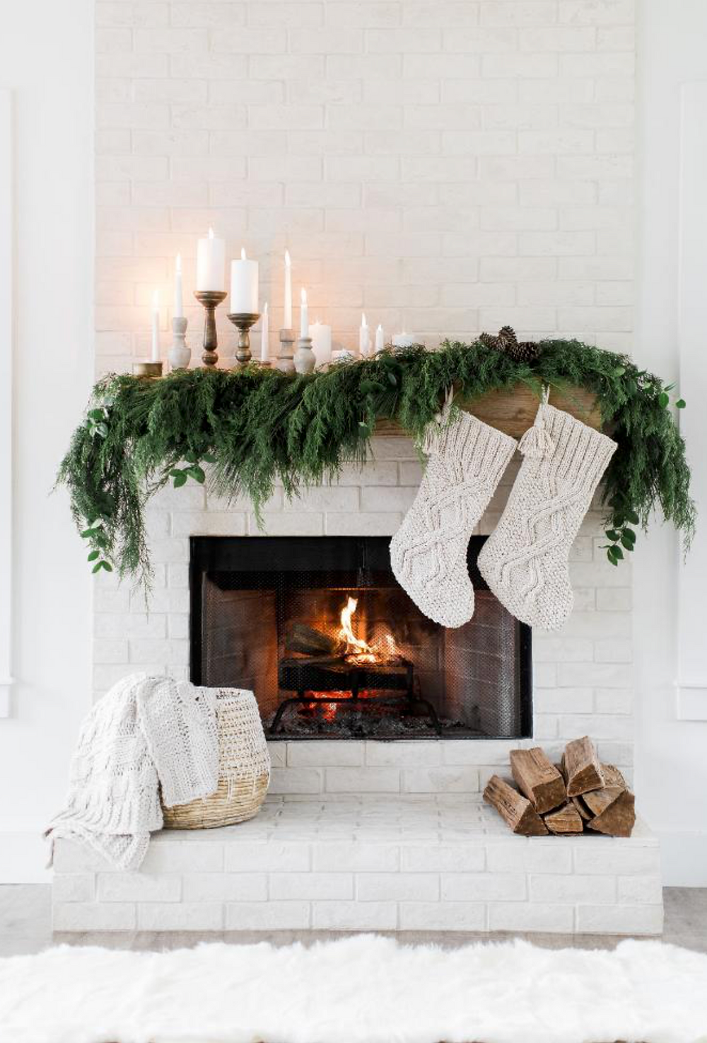 A white Christmas holiday fireplace mantel decorated with greenery, candles and a mirror with a basket of blankets for a modern and cozy feel.