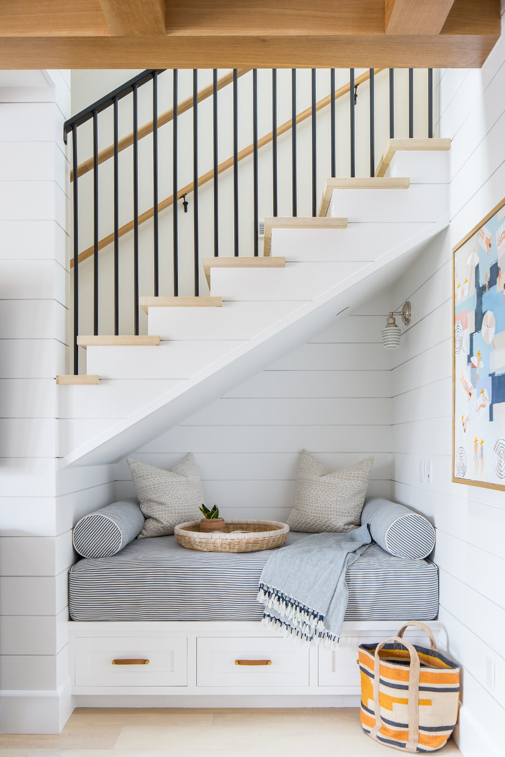 reading nook under stair way with three drawers grey foam mattress with throw pillow and blanket white shiplap detail stairs with wood treads black metal railing and hand rail woven basket abstract painting