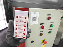 Plaqueta com QR-Code fixada na porta de um painel de comando, e uma mão com luva de trabalho segurando um celular que mostra a lista de documentos daquele ativo, com arquivos em PDF