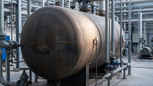 Vaso de pressão de aço carbono com costuras de solda e pátina, sob tubulação, em planta industrial, representando um ativo com longa história