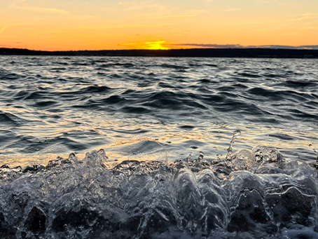 A close up shot of water reaching the shore with a light chop on the water and the last orange light of day setting in the distancet of day