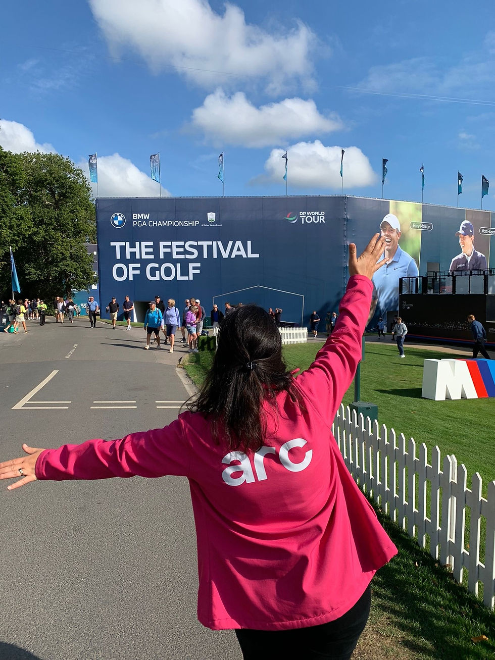 Woman in a pink jacket with "arc" on back raises arms at golf event. "THE FESTIVAL OF GOLF" banner and blue sky in background.