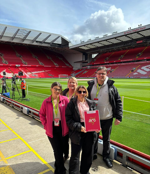 Four people standing on the sidelines of a football stadium. The stadium has red seats and a green field. The people are wearing lanyards with badges, and one person is holding a red folder with the text 'arc' on it. There are cameras and equipment set up in the background, ahead of the match