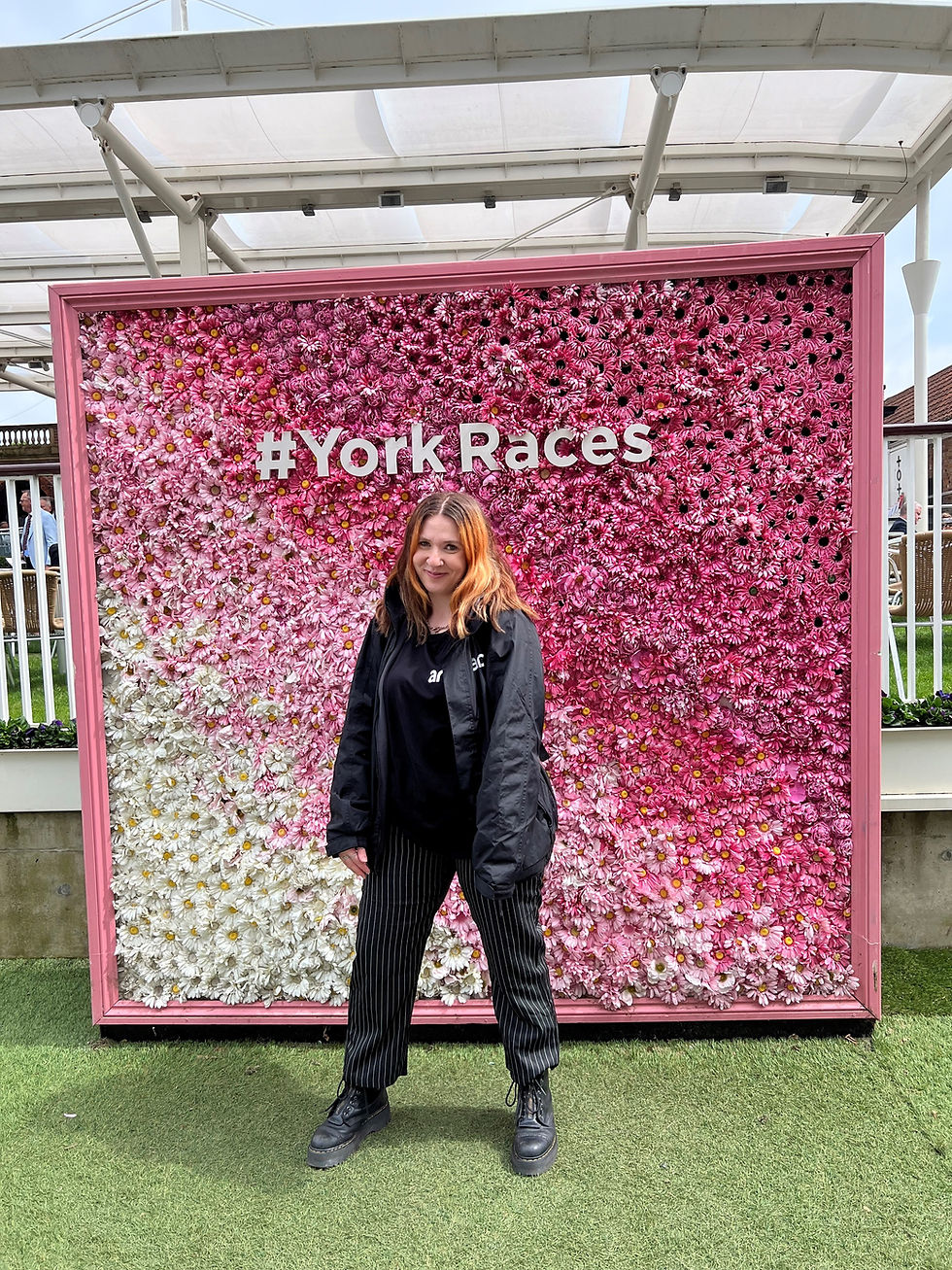 A person standing in front of a large floral wall display composed of flowers arranged in a gradient pattern, transitioning from white on the left to various shades of pink on the right. The floral wall has text that reads '#YorkRaces'. The person is wearing dark clothing and standing on green artificial grass.
