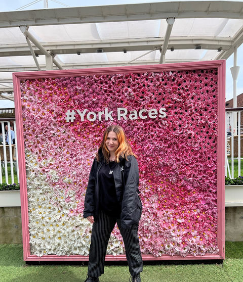 A person standing in front of a large floral wall display composed of flowers arranged in a gradient pattern, transitioning from white on the left to various shades of pink on the right. The floral wall has text that reads '#YorkRaces'. The person is wearing dark clothing and standing on green artificial grass.