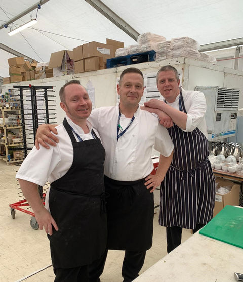 Three chefs in a kitchen or food preparation area, wearing chef uniforms including aprons. The background includes shelves with boxes and other kitchen supplies, a cart with trays, and various kitchen equipment.