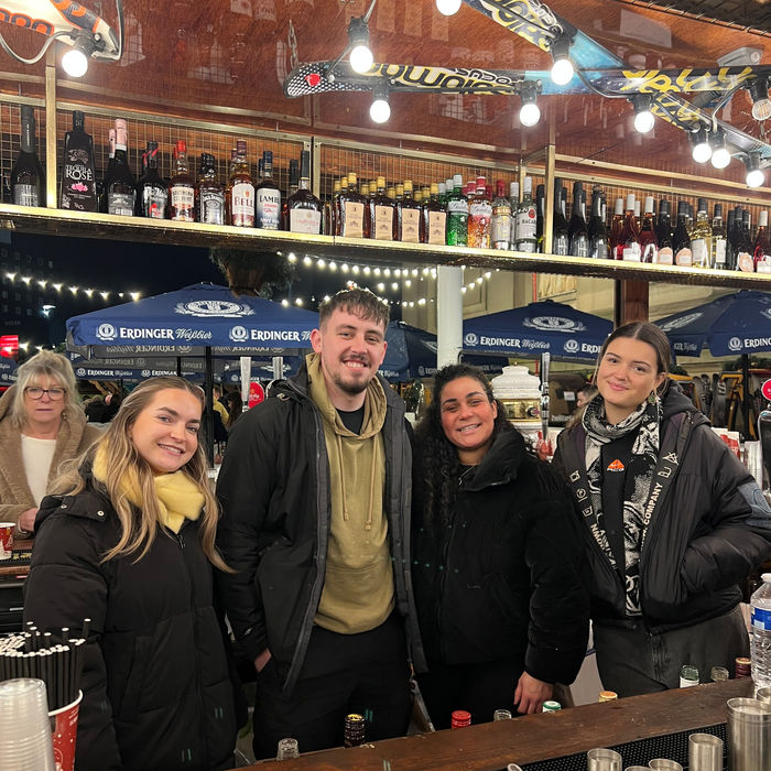 A group of four people, three women, one man wrapped up in winter coats stand behind a Christmas-themed bar.