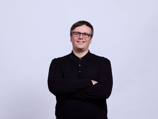 A man in a black long-sleeve top and black glasses smiles and crosses his arms