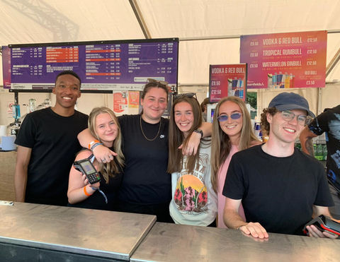 A group of six young adults smiling behind a festival bar counter inside a large tent. They are casually dressed, some with arms around each other, and appear to be working the bar. Behind them are colorful drink menus listing prices for various cocktails and beverages.