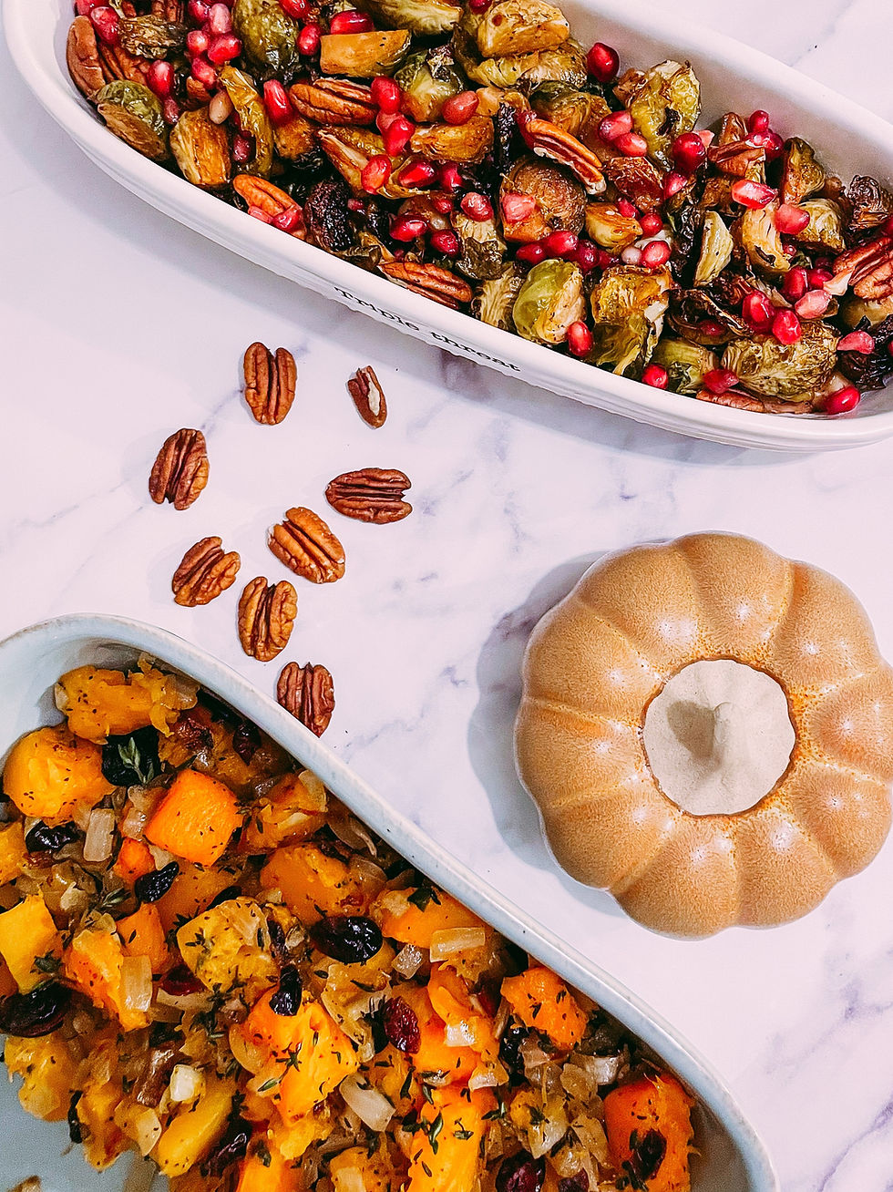 A table set for a holiday gathering with two brightly colored side dishes. One has roasted Brussels sprouts with pomegranate seeds and toasted pecans, while the other has orange butternut squash with dark red cranberries and caramelized onions.