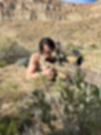 A female soldier aiming a sniper rifle, focused and poised, in a tactical outdoor environment.