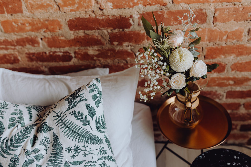 Green botanical-patterned pillows in front of white pillows beside a small table and brick wall.