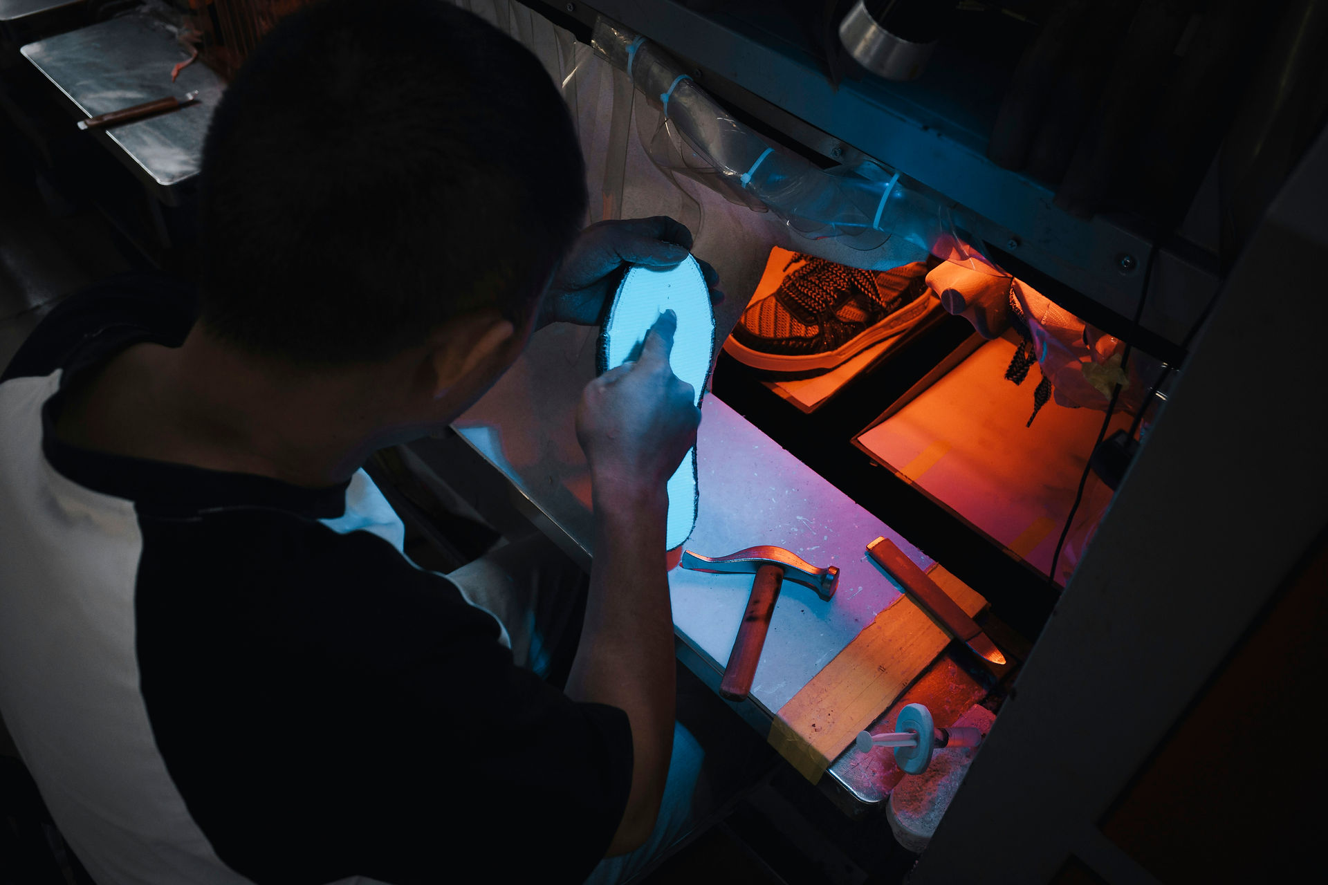 Ein Arbeiter hält an einem Arbeitsplatz eine Schuhsohle unter blaues Licht; in der Nähe liegen Schuhe und Handwerkzeuge.