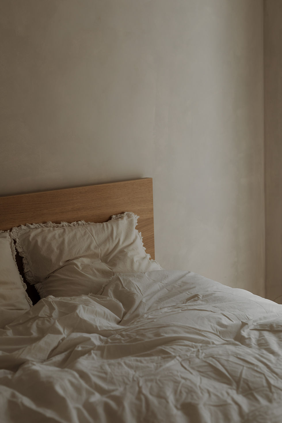 Rumpled light bedding and pillows on a bed with a simple wood headboard in a neutral room.