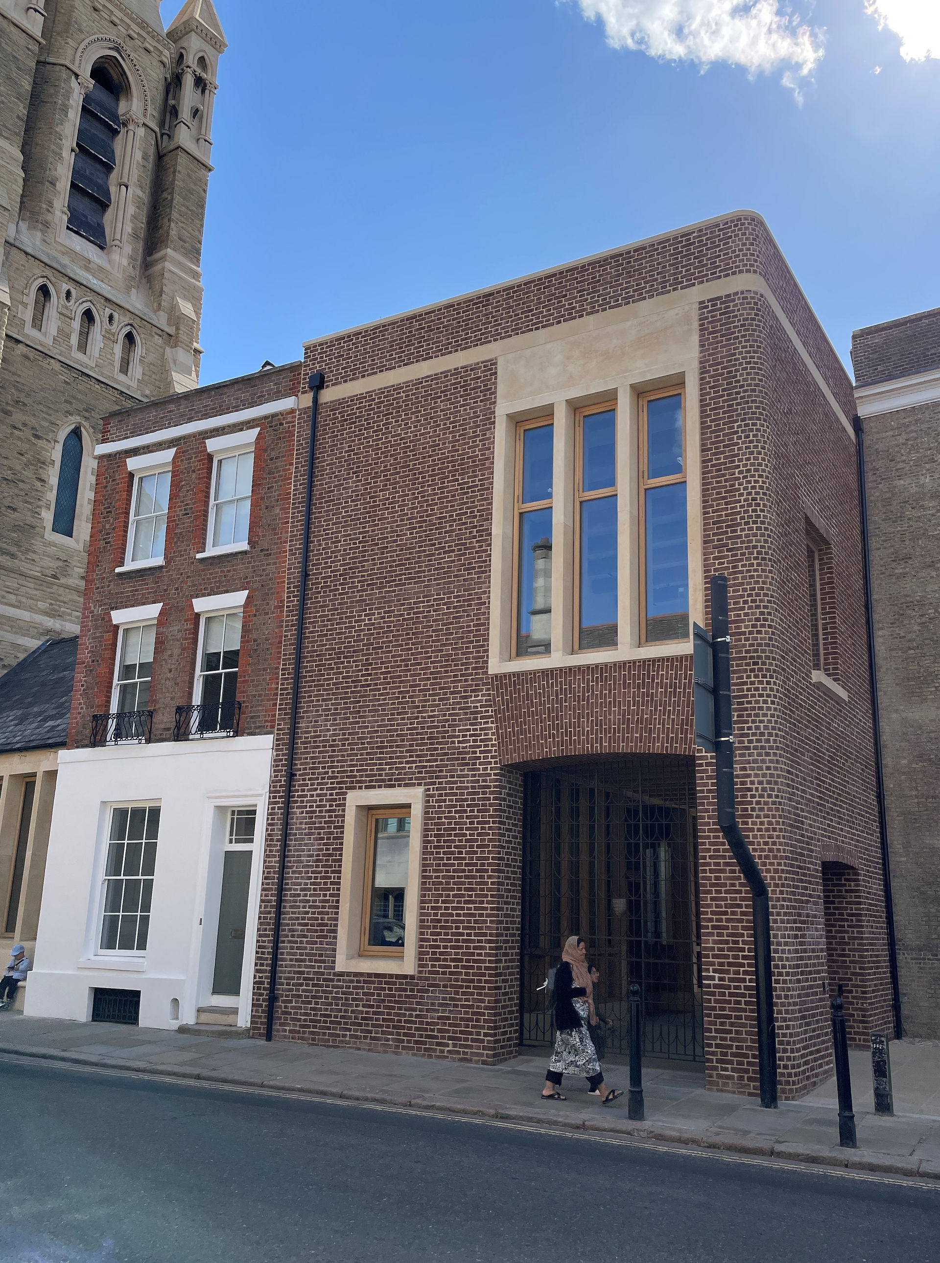 A newer brown-brick entrance building at Pembroke College’s Mill Lane redevelopment stands beside an older red-brick facade and church tower.