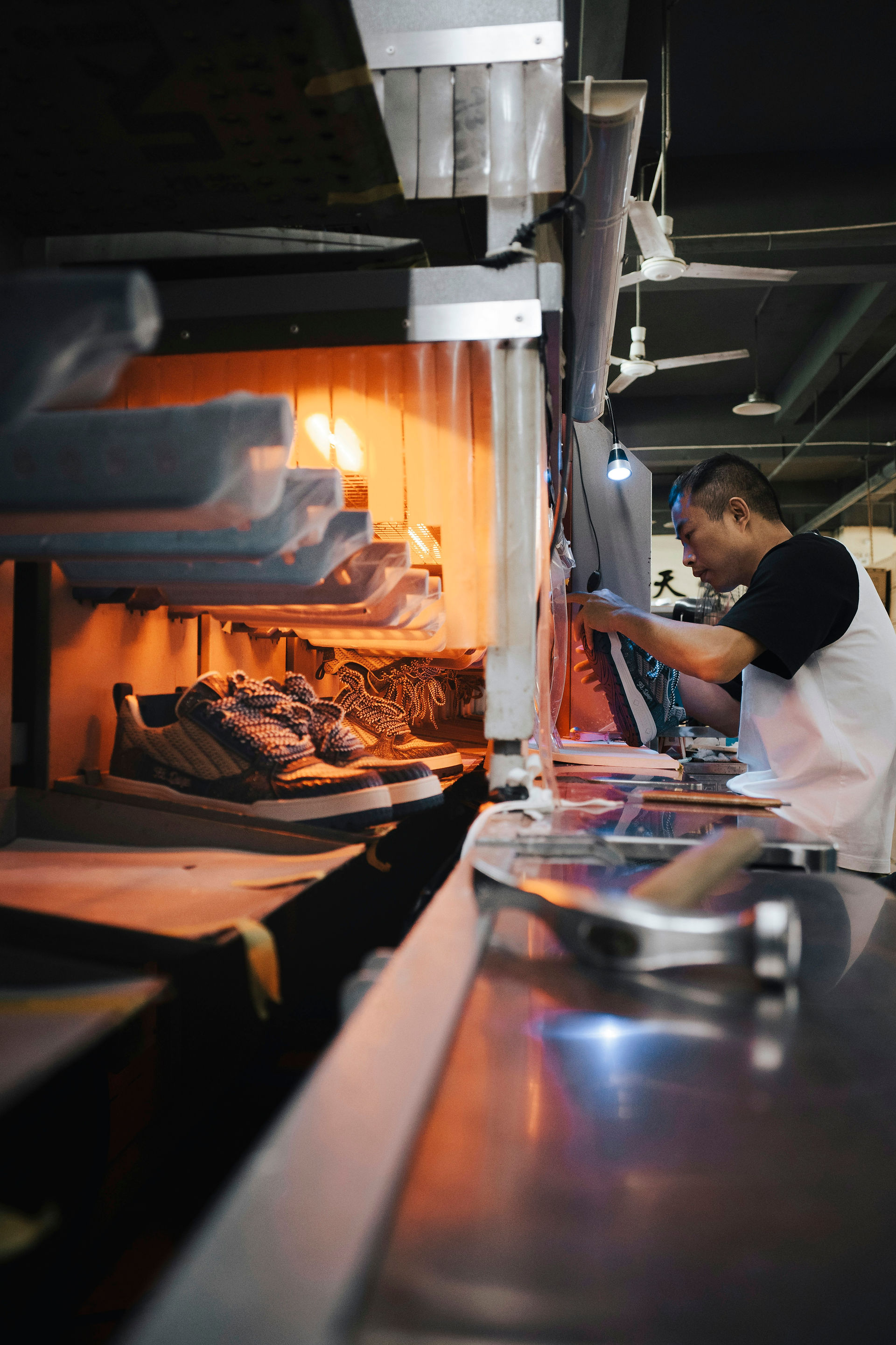 Ein Fabrikarbeiter handhabt einen Schuh neben einem orange beleuchteten Arbeitsplatz; weitere Schuhe sind in der Nähe angeordnet.
