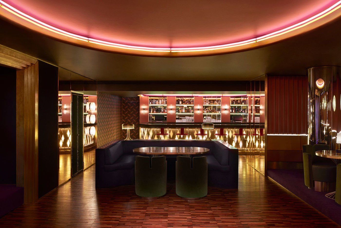 Wide room view connecting a blue central booth, tables, illuminated bar, mirrored walls and a curved ring of pink and amber light.