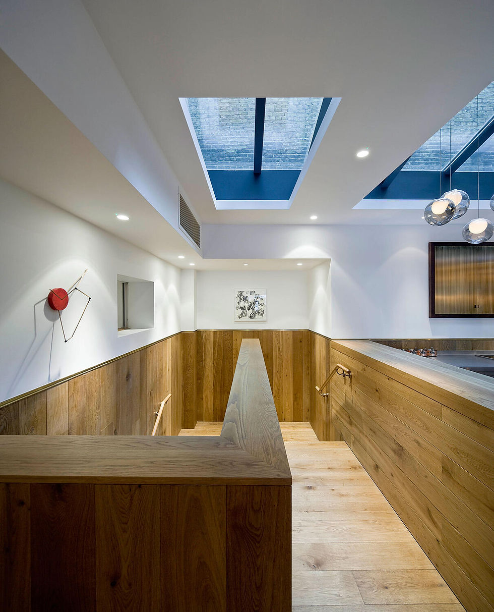 Rectangular ceiling openings and glass pendants appear above wooden stair balustrades and white walls.