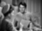 Man holding paper with sketches converses with a woman in a striped shirt in a room with cups and plates on the table, looking serious. Joel McCraw gawks at Jean Arthur eating an egg in the classic Hollywood romantic comedy film rom-com movie 1940s "The More the Merrier".