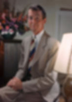 Man in a beige suit with a striped tie, sitting with hands clasped. Background has a vase of colorful flowers, creating a calm atmosphere. Color portrait photo of Hollywood movie star film actor middle aged older James Jimmy Stewart