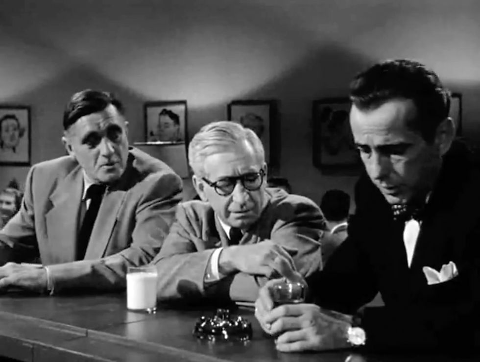 Three men in suits sit at a bar. One holds a glass, while another gestures. Black-and-white setting with framed art on walls. Serious mood. Morris Ankrum, Art Smith, Humphrey Bogart at the bar in the classic Hollywood movie romance Nicholas Ray film noir "In a Lonely Place"