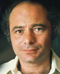 Man with short curly hair and a neutral expression, wearing a white shirt, set against a blurred dark background. Color porttrait photo of Hollywood movie star film TV actor younger Burt Young