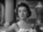 Black and white image of a woman with curly hair in a floral dress, sitting on a sofa in a classic room. She appears calm and thoughtful. Myrna Loy is the title role in the Hollywood film screwball 1930s comedy classic movie “Libeled Lady”