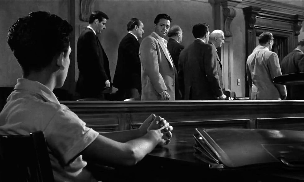 A seated boy watches a group of men in suits walk away in a courtroom. One man looks back. The setting is somber, in black and white. Edward Binns looks back at the accused as the jury leaves to deliberate in the classic movie Hollywood courtroom drama film "12 Angry Men" twelve