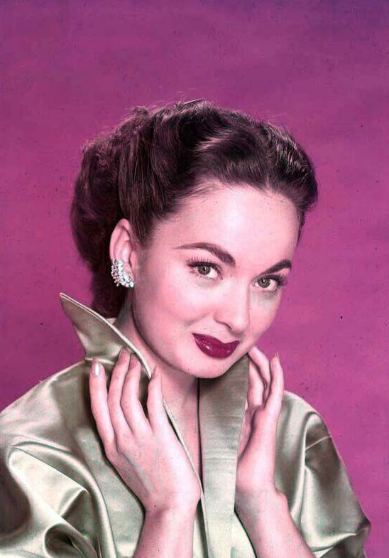Portrait of a smiling woman with dark hair, pearl earrings, and a satin coat against a pink background. Portrait photo of Hollywood movie star actress singer young Ann Blyth