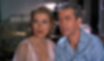 A woman in a pink dress and a man in a blue shirt gaze intently, appearing intrigued. Background shows a dimly lit room and chair. Grace Kelly and James Jimmy Stewart star in the Alfred Hitchcock classic Hollywood film suspense movie "Rear Window"