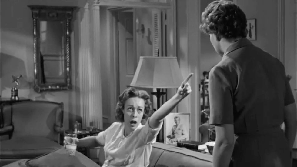 Woman on a couch points dramatically at a standing person, holding a glass. Elegant interior with a lamp and framed photo in the background. Black and white. Eileen Heckart drunk, points at Nancy Kelly in the cult classic film melodrama horror thriller Hollywood movie "The Bad Seed"