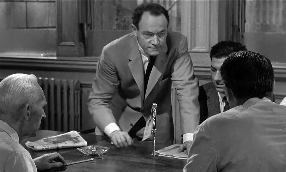 Black-and-white image of a man in a suit leaning on a table, intently discussing with seated men. Papers and an ashtray on the table, serious mood. E.G. Marshall with Joseph Sweeney, Jack Klugman, and Henry Fonda in the classic movie Hollywood courtroom drama film "12 Angry Men" twelve