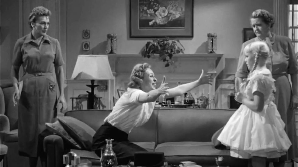 Woman reaches out to a smiling child in a living room, flanked by two standing women. Black and white scene with a cozy atmosphere. Nancy Kelly, Eileen Heckart, Patty McCormak, and Evelyn Varden in the cult classic film melodrama horror thriller Hollywood movie "The Bad Seed"