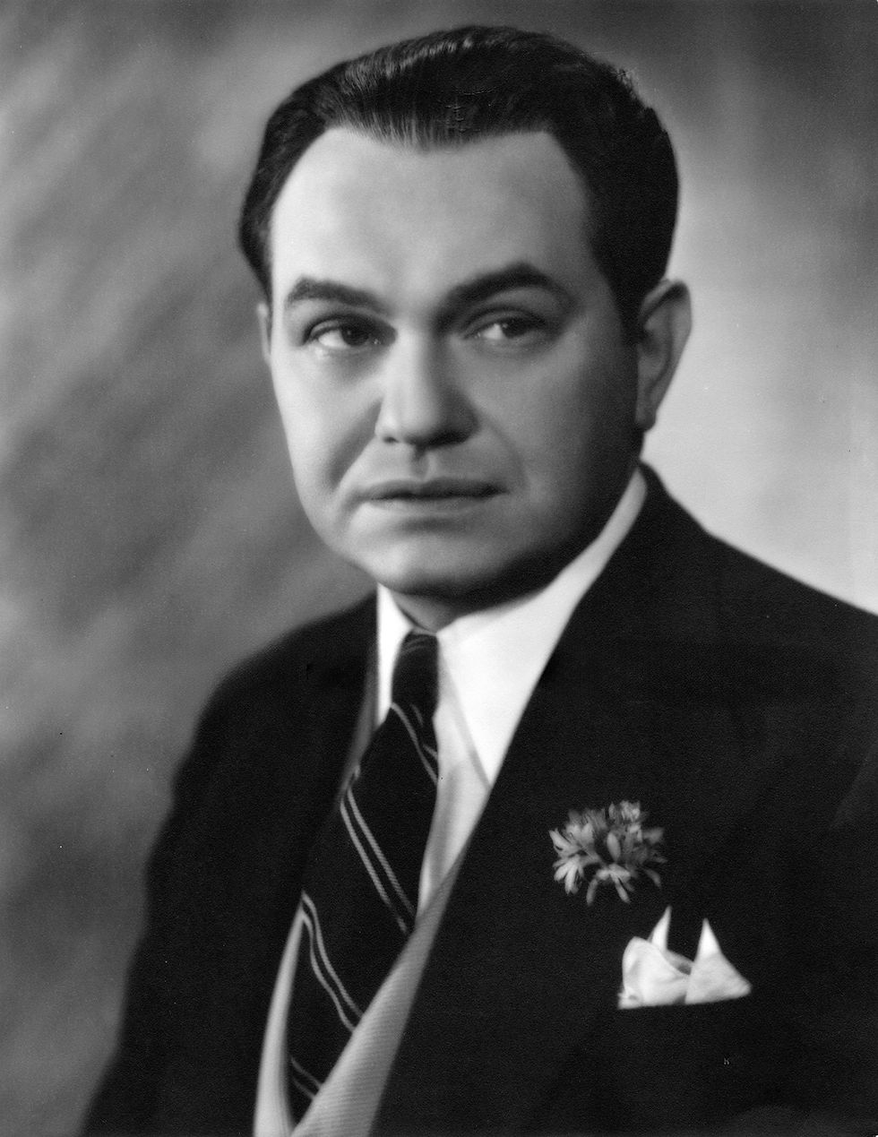Black and white portrait of a man in a suit with a striped tie. He has a neutral expression, flower on lapel, and blurred background. Portrait photo of Hollywood movie star film tough guy gangster actor young Edward G. Robinson