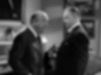 Two men in suits converse in a room; one gesturing with a cigar. Tense mood, framed art and clock visible in background. Black and white. William Frawley and Gene Lockhart in the classic movie Hollywood holiday Christmas Santa Claus film "Miracle on 34th Street"