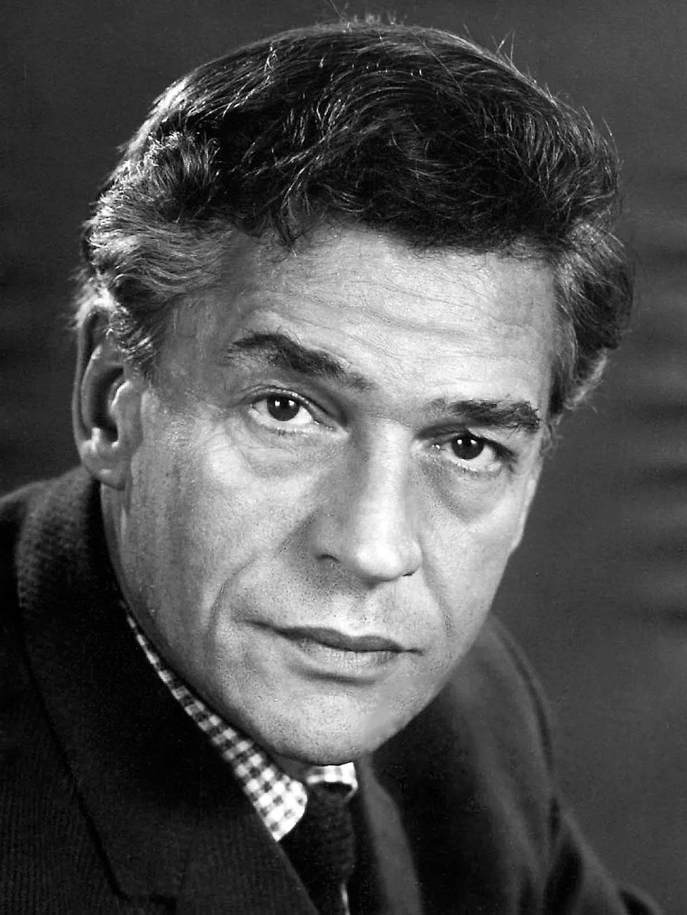 Black and white portrait of a man in a checkered shirt and jacket, looking intently at the camera. Calm expression, dark background. Portrait photo of British film and stage actor theater legend Hollywood movie star Paul Scofield