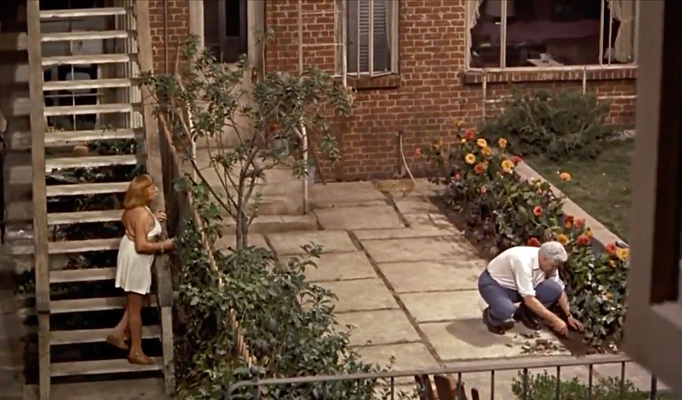 Woman in white dress stands by stairs, observing a man gardening on a patio. Red brick building and vibrant flowers in the background. Jesslyn Fax watches Raymond Burr dig in the courtyard's flowerbed in the Alfred Hitchcock classic Hollywood film suspense voyeuristic movie "Rear Window" Peeping Tom
