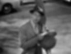 A man in a suit and hat leans on a lamp post, reading a newspaper. It's night on a city street; a parked car is visible. Black and white scene. Joel McCrea arrives with an airplane propeller in the classic Hollywood romantic comedy film rom-com movie 1940s "The More the Merrier".