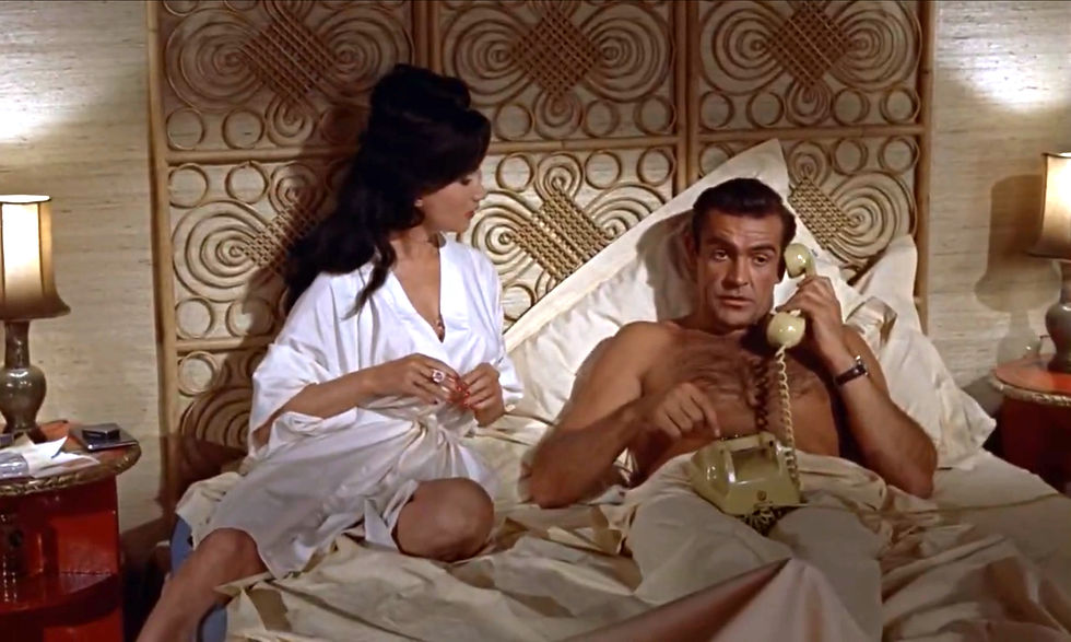 Woman in white robe sits on bed beside shirtless man using a beige corded phone in a warm, retro bedroom; both look serious. Zena Marshall and Sean Connery in the first classic James Bond movie espionage spy thriller 007 film 1962 "Dr. No"