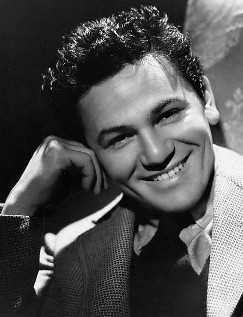 Smiling man in a suit leans on his hand, exuding a relaxed and confident mood in a softly lit environment. Black and white photo. Portrait photo of Hollywood movie actor film star tough guy young John Garfield
